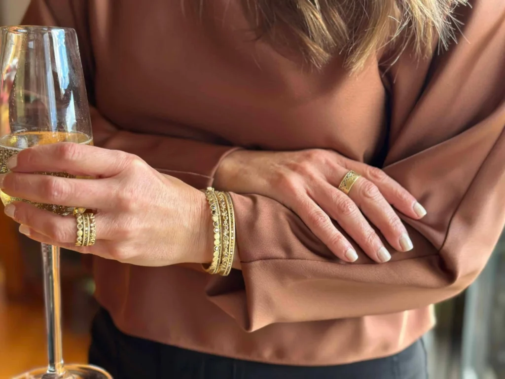 Woman holding a glass of champagne, wearing gold jewerly and a brown shirt.  